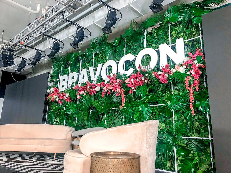 Greenery wall with dimensional Bravocon lettering and hanging pink flowers on a convention stage