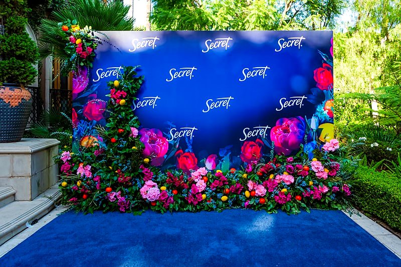 Blue step and repeat brand backdrop framed in pink, red and yellow flowers and citrus
