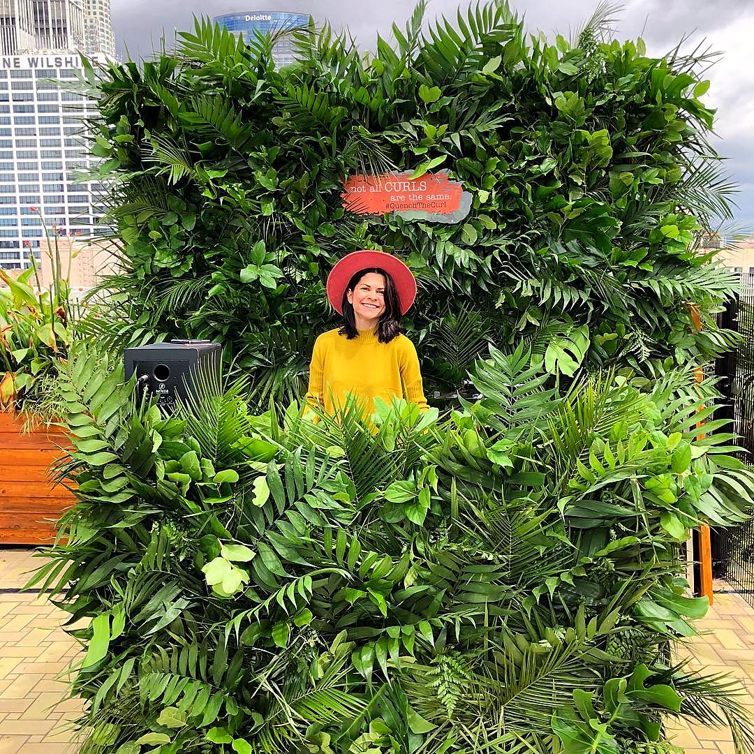 Carly Cylinder smiling behind a floor-to-ceiling greenery wall at an outdoor brand activation