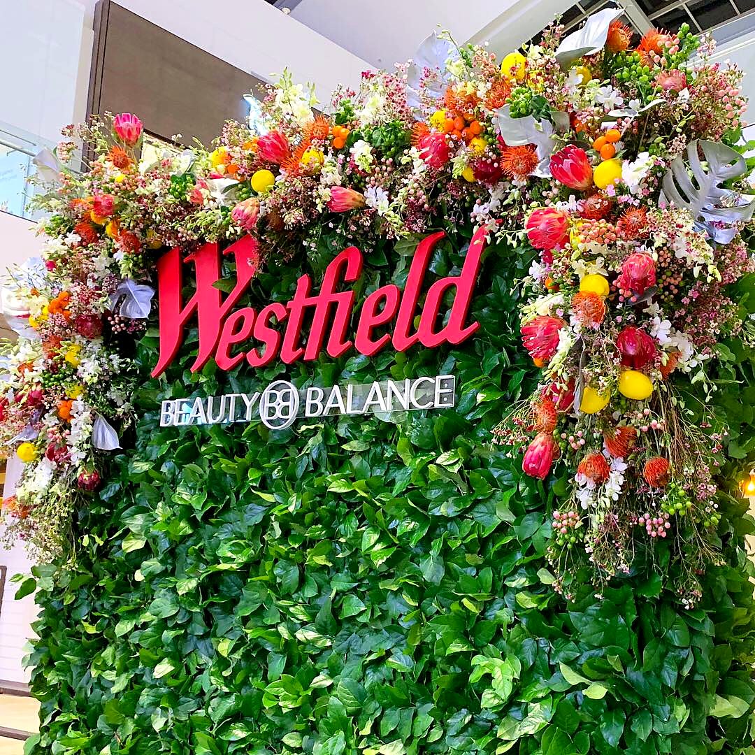 Greenery wall with a red logo and a crown of orange, pink and yellow flowers at a shopping center activation